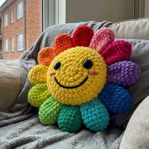 May include: A round, plush, crocheted pillow shaped like a flower with a bright yellow center featuring a black embroidered smile and two large black plastic eyes. The petals are arranged in a rainbow color sequence including red, pink, purple, blue, teal, green, lime, and orange.