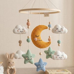 Könnte beinhalten: Ein Mobile für das Kinderzimmer mit einem runden Holzrahmen und hängenden Häkelelementen, darunter ein gelber Halbmond, weiße Wolken sowie mint- und blaufarbene Sterne. Jedes Teil ist mit einem gestickten, lächelnden Gesicht versehen.