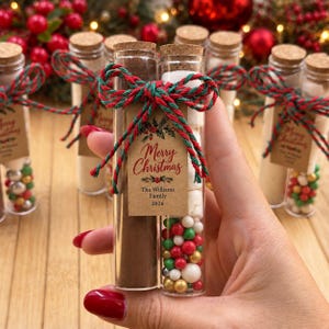 Op de afbeelding: Twee doorzichtige glazen reageerbuisjes met kurken stoppen, gevuld met bruin poeder en witte marshmallows met rode, groene en gouden strooisels. Een rood en groen touwtje houdt een beige label vast met de tekst Merry Christmas.