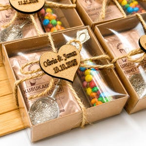 Può includere: Una scatola regalo rettangolare in cartone contenente una confezione di caffè marrone etichettata KURUKAHVECI MEHMET EFENDI, un tubo trasparente riempito con caramelle al cioccolato colorate e un cucchiaio in metallo argentato, legati con spago e un cuore in legno.
