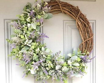 May include: A grapevine wreath decorated with faux lavender and eucalyptus sprigs. The wreath is hanging on a white door.