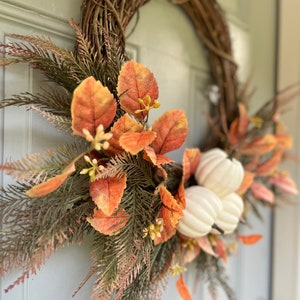 K&ouml;nnte beinhalten: Ein herbstliches Kranz mit einem geflochtenen Weinreben-Sockel. Er ist mit orangefarbenen und braunen Bl&auml;ttern, gr&uuml;nen Zweigen und kleinen wei&szlig;en K&uuml;rbissen verziert. Der Kranz ist ein dekoratives Element f&uuml;r eine T&uuml;r oder Wand.