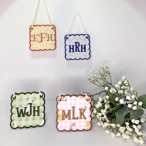 May include: Four decorative square signs with scalloped edges and white rope hangers. The signs feature embroidered initials and decorative patterns. Two signs have floral patterns, one with a duck and the other with a bunny. The other two signs have lace patterns.