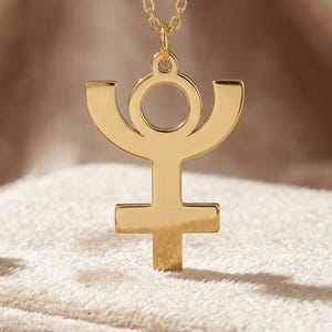 May include: A gold-toned necklace featuring a pendant shaped like the astrological symbol for Pluto. The pendant is suspended from a delicate matching gold-toned chain, showcasing a polished metallic finish and a clean, symmetrical design.