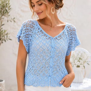 May include: A light blue crochet top featuring a V-neckline, short flutter sleeves, and a vertical row of matching blue buttons down the center front. The garment is crafted with an intricate openwork pattern throughout the bodice and sleeves.