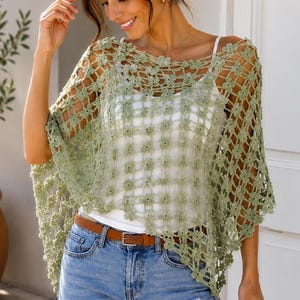May include: A sage green crochet poncho featuring a delicate floral pattern with an open-knit design. The garment is layered over a simple white camisole and paired with light blue denim jeans featuring a brown leather belt with a metal buckle.