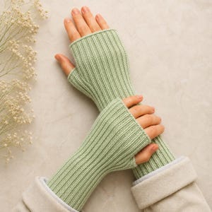 Peut inclure: Une paire de gants sans doigts vert sauge clair avec un motif en tricot côtelé vertical. Les gants dépassent le poignet et possèdent une ouverture dédiée pour le pouce, fabriqués dans un fil texturé doux avec un tricot uniforme.
