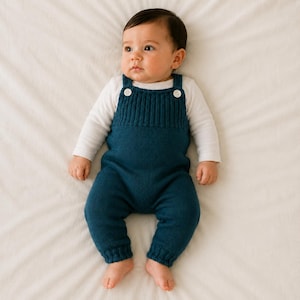 Op de afbeelding: Een baby draagt een wit shirt met lange mouwen onder een gebreide tuinbroek in de kleur blauwgroen. De tuinbroek heeft een geribbeld borstpaneel, twee witte knopen op de schouderbanden en geribbelde boorden bij de enkels voor een eenvoudig ontwerp.