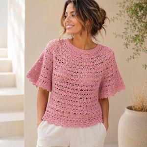 May include: A dusty rose crochet top featuring a round neckline, short flared sleeves, and a repeating openwork pattern throughout the garment. The piece is finished with a scalloped edge along the sleeves and the bottom hemline.