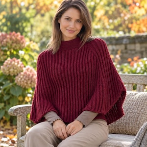 May include: A deep red, ribbed knit poncho featuring a high neckline and wide, flowing sleeves. The garment is crafted with a vertical textured pattern throughout, providing a structured yet comfortable fit that drapes over the shoulders and upper body.