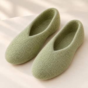 May include: A pair of sage green felted wool slippers with a rounded toe and a deep V-shaped opening at the top. The slippers feature a textured, fuzzy surface and a seamless construction, providing a simple slip-on design for indoor footwear.