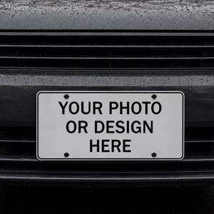 May include: A rectangular white license plate frame mounted on the front bumper of a dark gray car. The plate features the black text YOUR PHOTO OR DESIGN HERE centered in a bold sans-serif font, secured by four black screws at the corners.