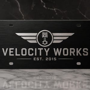 May include: Black license plate with the text "VELOCITY WORKS" in silver. The logo features a piston inside a circle with wings. The text "EST. 2015" is below the main text. The plate has a brushed metal texture.