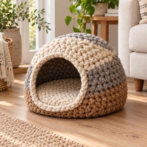 Può includere: Una cuccia per animali a forma di cupola realizzata in filato spesso e grosso con un motivo a blocchi di colore. Il design presenta fasce orizzontali color crema, grigio chiaro e marrone chiaro, con un'ampia apertura circolare frontale.