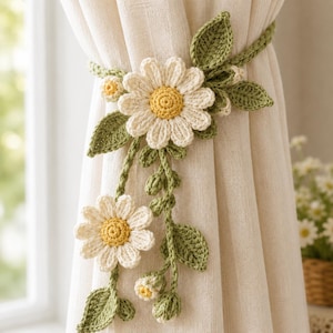 May include: A crochet curtain tieback featuring two large white daisy flowers with yellow centers, smaller daisy buds, and several green crocheted leaves attached to a long, thin green vine that wraps around a light beige fabric curtain.