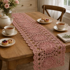 May include: A dusty rose crochet table runner featuring a repeating floral motif with an intricate openwork pattern. The runner is crafted from yarn and displays a scalloped edge, laid out flat to showcase the detailed circular stitching design.