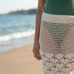 May include: A cream-colored crochet sarong featuring a grid-patterned upper section and a scalloped lace hem. The wrap-style skirt is secured at the side with ties adorned with wooden and turquoise-colored beads and matching fringe tassels.
