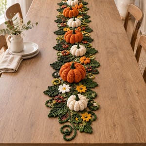 Può includere: Un lungo runner da tavola decorativo caratterizzato da un design all'uncinetto con zucche arancioni e bianche, foglie verde scuro e piccoli fiori gialli e bianchi. Il runner è disposto in linea centrale, mostrando trame di filato.