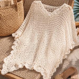 May include: A cream-colored crochet cover-up featuring an open-knit grid pattern, a V-neckline, and a decorative scalloped edge along the hem. The garment is crafted with a lightweight, airy texture and displays intricate stitching throughout the piece.