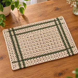 May include: A rectangular crochet placemat featuring an off-white openwork pattern with a dark green border design. The textile accessory has a grid-like structure with intersecting lines of green yarn forming a frame around the central area.