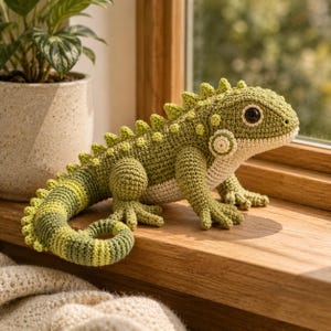Puede incluir: Un juguete de iguana tejido a ganchillo en tonos verde oliva y verde lima con el vientre color crema. El lagarto tiene el lomo texturizado con pequeñas espinas, una larga cola enroscada y grandes ojos de color marrón oscuro, con puntadas detalladas.