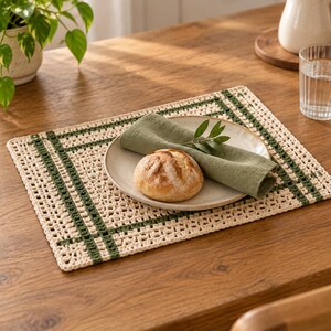 Op de afbeelding: Een rechthoekige gehaakte placemat met een gebroken witte basis en donkergroene rastervormige strepen. Op de placemat staat een rond keramisch bord met een knapperig broodje en een gevouwen olijfgroen linnen servet met een groen takje.