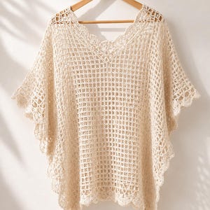 May include: A cream-colored crochet poncho featuring an open grid pattern, a V-neckline, and scalloped edges along the hem and sleeves. The garment is constructed with a lightweight, airy yarn and hangs from a wooden hanger, displaying a loose fit.