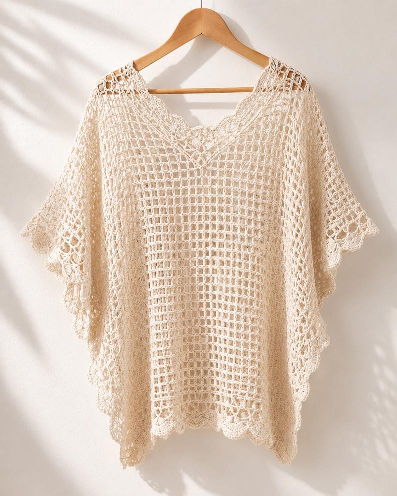 May include: A cream-colored crochet poncho featuring an open grid pattern, a V-neckline, and scalloped edges along the hem and sleeves. The garment is constructed with a lightweight, airy yarn and hangs from a wooden hanger, displaying a loose fit.