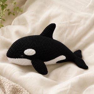 May include: A handcrafted crochet orca toy featuring a black and white colour scheme. The plush figure displays the characteristic dorsal fin, side flippers, and tail fluke of an orca, with a white patch near the eye and a white underside.