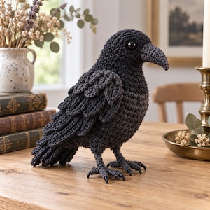 May include: A handcrafted crochet crow figurine made with dark charcoal yarn. The bird features a textured plumage design with layered wing feathers, a curved beak, and two small black eyes. It stands on two crocheted legs with clawed feet.