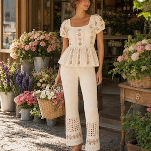 May include: A cream-colored crochet top and pants set with floral embroidery. The top has a square neckline and short sleeves. The pants have a flared leg with a decorative floral pattern at the hem. A woven basket filled with flowers is held in the hand.