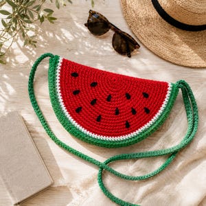 May include: A handmade, crochet watermelon-shaped crossbody bag. The bag features a red body with black seed details, a white rind, and a green border. The strap is green. Sunglasses and a straw hat are in the background.