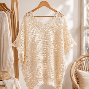 May include: A cream-colored crochet poncho featuring an open-knit grid pattern throughout the body, a V-neckline, and scalloped edges along the hem and sleeves. The garment is displayed on a wooden hanger, showcasing its intricate, lightweight texture.