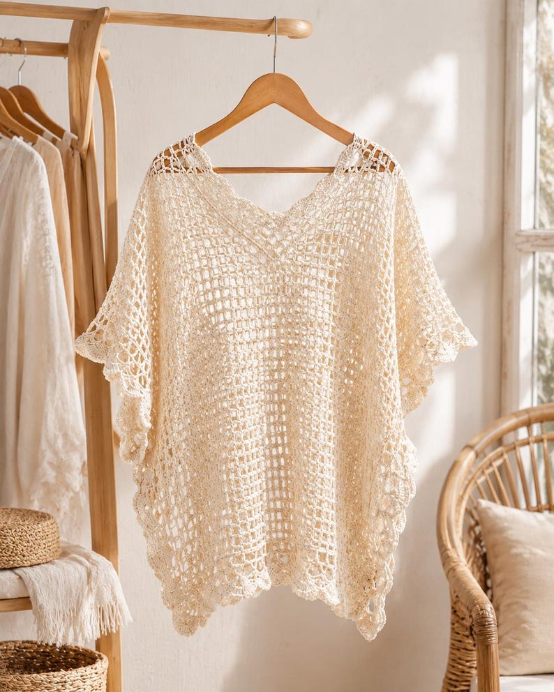 May include: A cream-colored crochet poncho featuring an open-knit grid pattern throughout the body, a V-neckline, and scalloped edges along the hem and sleeves. The garment is displayed on a wooden hanger, showcasing its intricate, lightweight texture.
