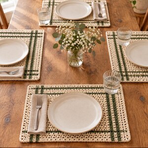 Op de afbeelding: Een set rechthoekige gehaakte placemats met een cr&egrave;mekleurige basis en donkergroene strepen in ruitpatroon. Elke placemat is op een houten tafel gerangschikt met een wit keramisch bord, een beige linnen servet, een zilveren vork en een glas.