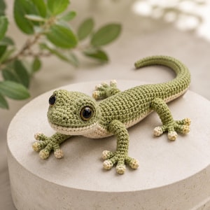 May include: A crocheted lizard figurine featuring a textured olive green body with a cream-coloured underbelly, large dark brown eyes, and small cream-coloured tips on each of its toes. The lizard is posed with its long tail curved to the side.