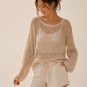 Può includere: Una donna indossa un maglione beige a maglia aperta con maniche lunghe e scollo tondo. Indossa anche una canotta beige chiaro e pantaloncini abbinati con coulisse in vita. L'outfit è casual e confortevole.