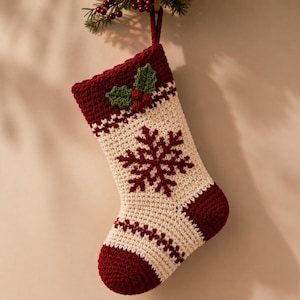 May include: A cream-colored crocheted Christmas stocking featuring a dark red cuff and toe. The cuff is decorated with green crocheted holly leaves and red berries. A large dark red snowflake motif is centered on the cream-colored body.