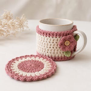 May include: A ceramic mug featuring a cream-colored body and a crocheted cozy in dusty rose and cream yarn. The cozy includes a decorative crocheted flower with a small wooden button center and green leaves. A matching circular crochet coaster.