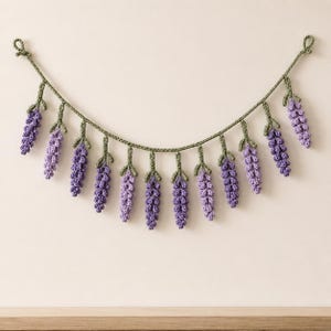 Può includere: Una ghirlanda decorativa con dodici rametti di lavanda realizzati all'uncinetto in tonalità viola, ciascuno fissato a un cordoncino centrale verde oliva. I rametti pendono verticalmente, creando una disposizione drappeggiata a festone.