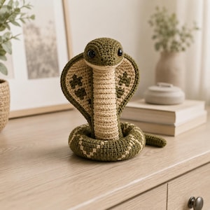 Könnte beinhalten: Ein gehäkeltes Kobra-Stofftier mit geringeltem Körper und aufgerichteter Haube. Die Schlange ist aus olivgrünem und cremefarbenem Garn gefertigt und hat dunkelbraune Kunststoffaugen sowie ein strukturiertes Rippenmuster an Hals und Haube.