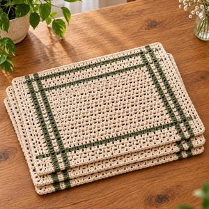 Op de afbeelding: Een stapel van drie rechthoekige gehaakte placemats met een cr&egrave;mekleurige basis en een rasterpatroon van donker bosgroene strepen. De placemats hebben een getextureerde, open geweven structuur met een consistent randontwerp op elk stuk.