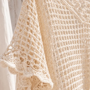 May include: An off-white crochet top featuring an open diamond-shaped mesh pattern throughout the body. The garment has wide, flared sleeves with a scalloped edge detail and a V-neckline finished with a matching scalloped crochet trim.