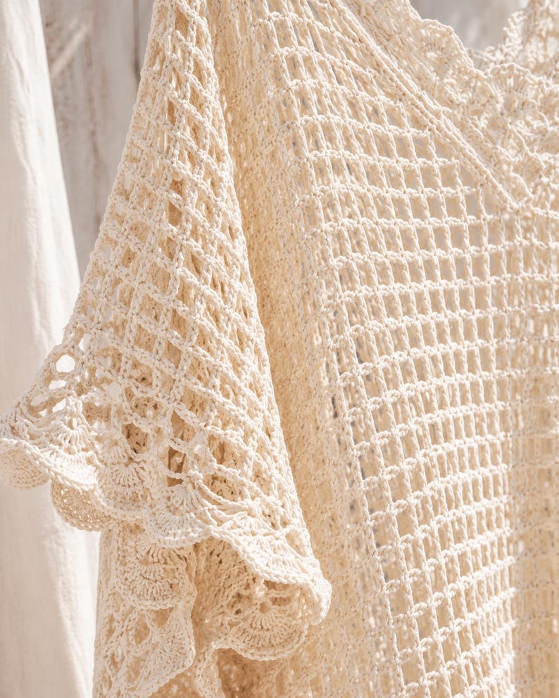 May include: An off-white crochet top featuring an open diamond-shaped mesh pattern throughout the body. The garment has wide, flared sleeves with a scalloped edge detail and a V-neckline finished with a matching scalloped crochet trim.