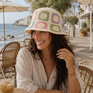 May include: A crochet bucket hat featuring a pattern of square motifs in pastel shades of pink, green, blue, and lavender. Each square contains a central white daisy design with a yellow center, all connected by a cream-colored yarn border.
