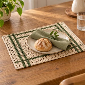 Op de afbeelding: Een rechthoekige gehaakte placemat met een gebroken witte basis en donkergroene rastervormige strepen. Op de placemat staat een rond keramisch bord met een knapperig broodje en een gevouwen olijfgroen linnen servet met een groen takje.