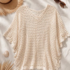 May include: A cream-colored crochet top featuring a V-neckline, short wide sleeves, and a grid-like openwork pattern. The garment is finished with a scalloped edge along the sleeves and hemline, providing a textured and lightweight appearance.