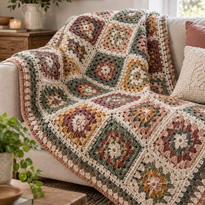 May include: A crochet blanket featuring a repeating granny square pattern in a muted color palette of sage green, dusty rose, mustard yellow, and cream. The squares are joined together with cream yarn, creating a textured, geometric design.