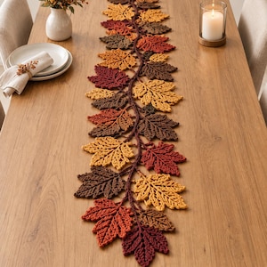 May include: A long, decorative table runner crafted from crocheted maple leaves in shades of deep burgundy, burnt orange, golden yellow, and dark brown. The leaves are arranged in a dense, overlapping vine-like pattern that runs down the centre.