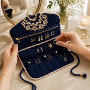 May include: A navy blue crochet jewelry organizer featuring a cream-colored lace trim on the flap, a zippered compartment, multiple earring holes, ring slots, and snap-button tabs for necklaces, secured with long braided ties and a gold-toned bead.